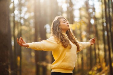 Bir kadın, sonbaharda yapraklar ve uzun ağaçlarla çevrili güneşli bir ormanda yoga yapıyor. Heather yaşam tarzı.
