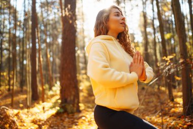 Bir kadın, sonbaharda yapraklar ve uzun ağaçlarla çevrili güneşli bir ormanda yoga yapıyor. Heather yaşam tarzı.