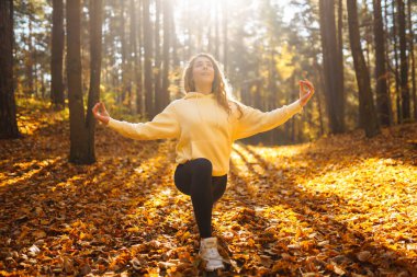 Bir kadın, sonbaharda yapraklar ve uzun ağaçlarla çevrili güneşli bir ormanda yoga yapıyor. Heather yaşam tarzı.