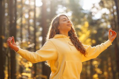 Bir kadın, sonbaharda yapraklar ve uzun ağaçlarla çevrili güneşli bir ormanda yoga yapıyor. Heather yaşam tarzı.