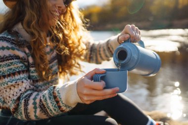 Genç bir kadın, güneşli bir sonbahar öğleden sonrasında, renkli yapraklar ve ışıldayan suyla çevrili nehir kenarında sıcak bir içkinin tadını çıkarıyor. Seyahat konsepti, turist, rahatla..