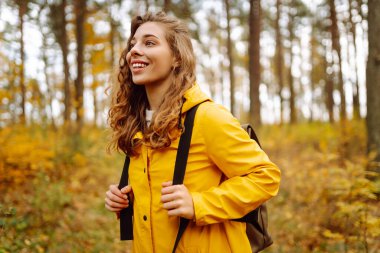 Sarı yağmurluklu genç bir kadın güneşli bir öğleden sonra yürüyüşü sırasında renkli yeşilliklerle dolu canlı bir sonbahar ormanını keşfediyor. Sonbahar manzarası.