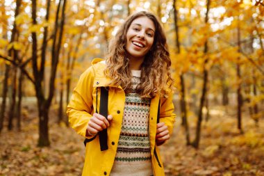 Sarı yağmurluklu genç bir kadın güneşli bir öğleden sonra yürüyüşü sırasında renkli yeşilliklerle dolu canlı bir sonbahar ormanını keşfediyor. Sonbahar manzarası.
