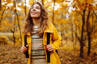 Sarı yağmurluklu genç bir kadın güneşli bir öğleden sonra yürüyüşü sırasında renkli yeşilliklerle dolu canlı bir sonbahar ormanını keşfediyor. Sonbahar manzarası.