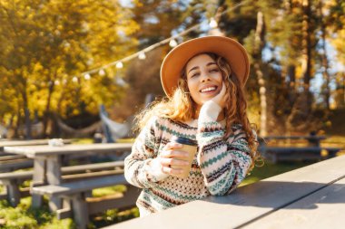 Neşeli bir kadın, göl kenarında sonbahar ağaçlarıyla çevrili sıcacık bir kazakla dışarıda sıcak bir içecekten keyif alıyor. Seyahatin, tatilin ve sıcak kahvenin tadını çıkar.. 