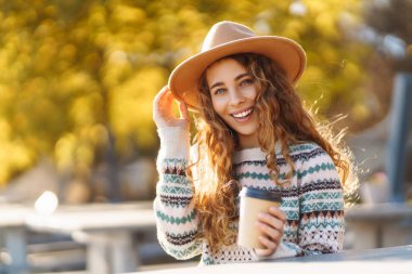 Neşeli bir kadın, göl kenarında sonbahar ağaçlarıyla çevrili sıcacık bir kazakla dışarıda sıcak bir içecekten keyif alıyor. Seyahatin, tatilin ve sıcak kahvenin tadını çıkar.. 