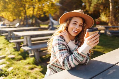 Neşeli bir kadın, göl kenarında sonbahar ağaçlarıyla çevrili sıcacık bir kazakla dışarıda sıcak bir içecekten keyif alıyor. Seyahatin, tatilin ve sıcak kahvenin tadını çıkar.. 