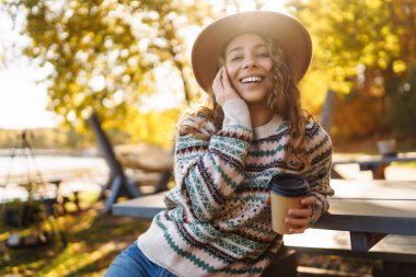 Neşeli bir kadın, göl kenarında sonbahar ağaçlarıyla çevrili sıcacık bir kazakla dışarıda sıcak bir içecekten keyif alıyor. Seyahatin, tatilin ve sıcak kahvenin tadını çıkar.. 