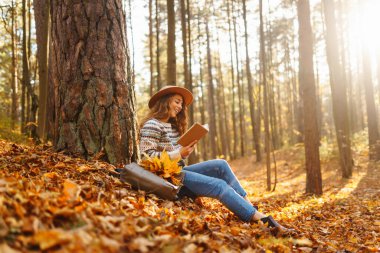 Bir kadın, düşen yaprakların üzerinde bisikletinin yanında otururken, sonbahar boyunca huzurlu bir ormanda kitap okumaktan hoşlanır.