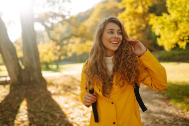 Sarı ceketli genç bir kadın güneşli bir sonbahar gününün tadını çıkarırken renkli sonbahar yapraklarıyla süslenmiş bir parkta yürüyor. Rahatlama, keyif alma, doğayla baş başa kalma..