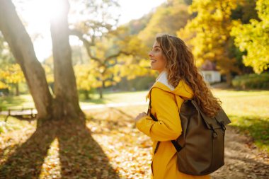 Sarı ceketli genç bir kadın güneşli bir sonbahar gününün tadını çıkarırken renkli sonbahar yapraklarıyla süslenmiş bir parkta yürüyor. Rahatlama, keyif alma, doğayla baş başa kalma..