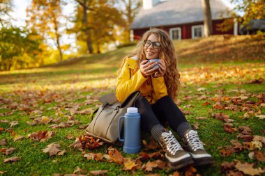 Sarı ceketli neşeli kadın renkli sonbahar yapraklarının arasında oturur, dışarıda huzurlu bir anın tadını çıkarırken sıcak bir içeceği bardağa doldurur. Sonbahar manzarası.