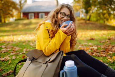 Sarı ceketli neşeli kadın renkli sonbahar yapraklarının arasında oturur, dışarıda huzurlu bir anın tadını çıkarırken sıcak bir içeceği bardağa doldurur. Sonbahar manzarası.