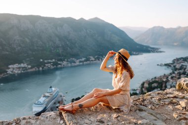 Gün batımında Karadağ 'da Kotor Körfezi' nin çarpıcı manzarasının keyfini çıkaran bir kadın, suyun yakınındaki kayalık bir çıkıntıda oturuyor. Turizm, rahatlama, gençlik ve doğa kavramı.