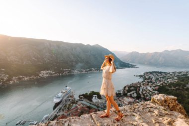 Gün batımında Karadağ 'da Kotor Körfezi' nin çarpıcı manzarasının keyfini çıkaran bir kadın, suyun yakınındaki kayalık bir çıkıntıda oturuyor. Turizm, rahatlama, gençlik ve doğa kavramı.