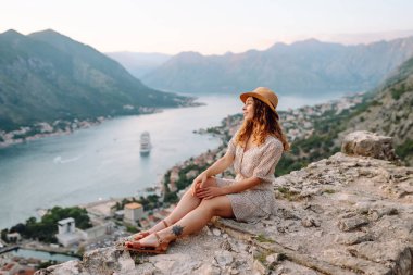 Gün batımında Karadağ 'da Kotor Körfezi' nin çarpıcı manzarasının keyfini çıkaran bir kadın, suyun yakınındaki kayalık bir çıkıntıda oturuyor. Turizm, rahatlama, gençlik ve doğa kavramı.