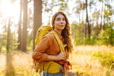Kıvırcık saçlı genç bir kadın, öğleden sonra güneşli bir ormanda omuzlarında sarı bir sırt çantası taşıyarak sakin bir yürüyüş yapıyor. Turizm, rahatlama, gençlik ve doğa kavramı.