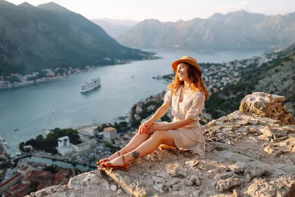 Gün batımında Karadağ 'da Kotor Körfezi' nin çarpıcı manzarasının keyfini çıkaran bir kadın, suyun yakınındaki kayalık bir çıkıntıda oturuyor. Turizm, rahatlama, gençlik ve doğa kavramı.