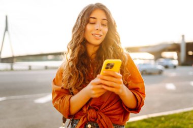 Açık havada güneşli bir günün tadını çıkaran genç bir kadın gün batımında şehrin rıhtımında akıllı telefonunu kullanıyor. Teknoloji, bağlantı, blog yazma, modern yaşam