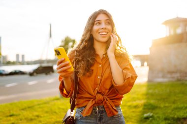 Açık havada güneşli bir günün tadını çıkaran genç bir kadın gün batımında şehrin rıhtımında akıllı telefonunu kullanıyor. Teknoloji, bağlantı, blog yazma, modern yaşam