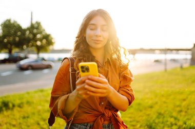 Açık havada güneşli bir günün tadını çıkaran genç bir kadın gün batımında şehrin rıhtımında akıllı telefonunu kullanıyor. Teknoloji, bağlantı, blog yazma, modern yaşam