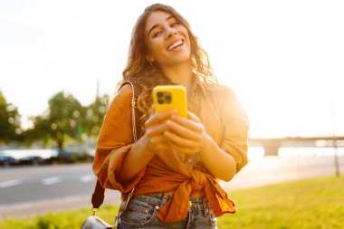 Açık havada güneşli bir günün tadını çıkaran genç bir kadın gün batımında şehrin rıhtımında akıllı telefonunu kullanıyor. Teknoloji, bağlantı, blog yazma, modern yaşam
