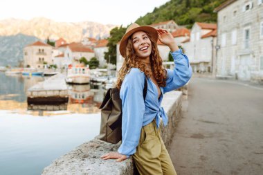 Neşeli bir kadın, sakin dağlar ve teknelerle çevrili bir şapka ve açık mavi bluz giyen büyüleyici bir sahil kasabasında deniz kenarında poz veriyor. Avrupa seyahati.