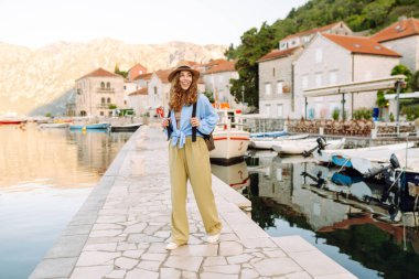 Neşeli bir kadın, sakin dağlar ve teknelerle çevrili bir şapka ve açık mavi bluz giyen büyüleyici bir sahil kasabasında deniz kenarında poz veriyor. Avrupa seyahati.