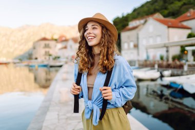 Neşeli bir kadın, sakin dağlar ve teknelerle çevrili bir şapka ve açık mavi bluz giyen büyüleyici bir sahil kasabasında deniz kenarında poz veriyor. Avrupa seyahati.
