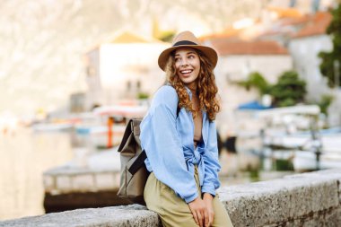 Neşeli bir kadın, sakin dağlar ve teknelerle çevrili bir şapka ve açık mavi bluz giyen büyüleyici bir sahil kasabasında deniz kenarında poz veriyor. Avrupa seyahati.