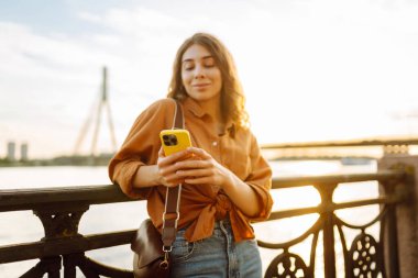 Gün batımında rıhtım kenarında akıllı telefon kullanan genç bir kadın, sıcak akşam ışıklarının keyfini çıkarırken günlük kıyafetler giyiyor. Yaşam tarzı, seyahat, doğa, aktif yaşam.