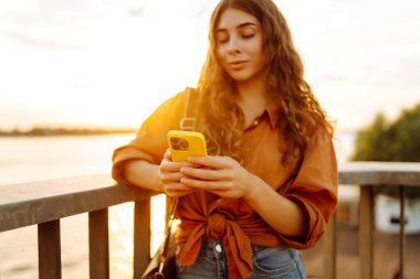 Young woman using smartphone by the waterfront during sunset, wearing a casual outfit while enjoying the warm evening light. Lifestyle, travel, nature, active life. clipart