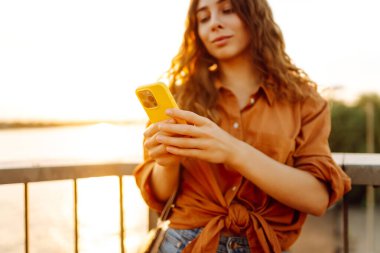 Gün batımında rıhtım kenarında akıllı telefon kullanan genç bir kadın, sıcak akşam ışıklarının keyfini çıkarırken günlük kıyafetler giyiyor. Yaşam tarzı, seyahat, doğa, aktif yaşam.