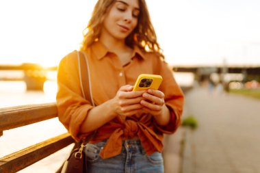 Gün batımında rıhtım kenarında akıllı telefon kullanan genç bir kadın, sıcak akşam ışıklarının keyfini çıkarırken günlük kıyafetler giyiyor. Yaşam tarzı, seyahat, doğa, aktif yaşam.