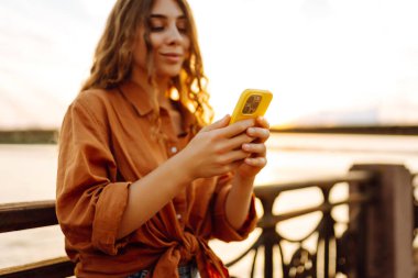 Gün batımında rıhtım kenarında akıllı telefon kullanan genç bir kadın, sıcak akşam ışıklarının keyfini çıkarırken günlük kıyafetler giyiyor. Yaşam tarzı, seyahat, doğa, aktif yaşam.