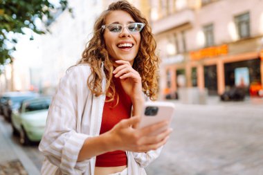 Güneşli bir günün tadını çıkaran genç bir kadın şık bir güneş gözlüğü ve neşeli bir ifadeyle canlı bir şehir ortamında telefonunu kullanıyor. Seyahat, blog, hafta sonu, rahatlama ve yaşam tarzı konsepti.