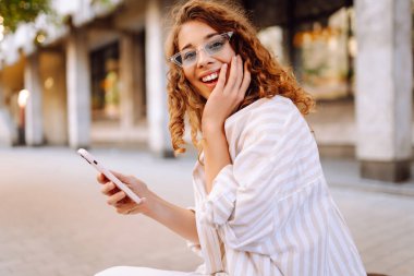 Güneşli bir günün tadını çıkaran genç bir kadın şık bir güneş gözlüğü ve neşeli bir ifadeyle canlı bir şehir ortamında telefonunu kullanıyor. Seyahat, blog, hafta sonu, rahatlama ve yaşam tarzı konsepti.
