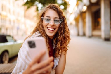 Güneşli bir günün tadını çıkaran genç bir kadın şık bir güneş gözlüğü ve neşeli bir ifadeyle canlı bir şehir ortamında telefonunu kullanıyor. Seyahat, blog, hafta sonu, rahatlama ve yaşam tarzı konsepti.