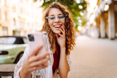 Güneşli bir günün tadını çıkaran genç bir kadın şık bir güneş gözlüğü ve neşeli bir ifadeyle canlı bir şehir ortamında telefonunu kullanıyor. Seyahat, blog, hafta sonu, rahatlama ve yaşam tarzı konsepti.