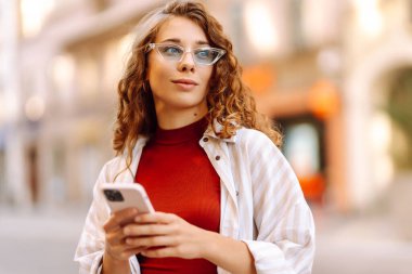 Güneşli bir günün tadını çıkaran genç bir kadın şık bir güneş gözlüğü ve neşeli bir ifadeyle canlı bir şehir ortamında telefonunu kullanıyor. Seyahat, blog, hafta sonu, rahatlama ve yaşam tarzı konsepti.