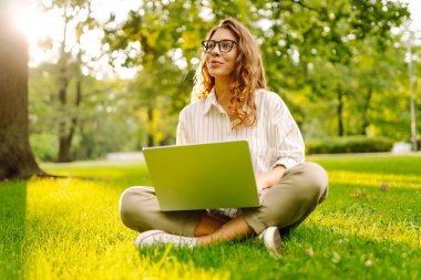 Kıvırcık saçlı genç bir kadın dışarıda bilgisayarını kullanarak çimlerin üzerinde rahatça oturuyor. İş, serbest meslek, online eğitim.