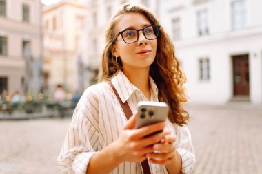 Kıvırcık saçlı ve gözlüklü genç bir kadın gece şehir meydanında akıllı telefonunu incelikle kullanıyor. Arkadaşlarla iletişim. Teknoloji, bağlantı, blog yazma, modern yaşam tarzı kavramı