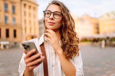 Kıvırcık saçlı ve gözlüklü genç bir kadın gece şehir meydanında akıllı telefonunu incelikle kullanıyor. Arkadaşlarla iletişim. Teknoloji, bağlantı, blog yazma, modern yaşam tarzı kavramı