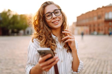 Kıvırcık saçlı ve gözlüklü genç bir kadın gece şehir meydanında akıllı telefonunu incelikle kullanıyor. Arkadaşlarla iletişim. Teknoloji, bağlantı, blog yazma, modern yaşam tarzı kavramı