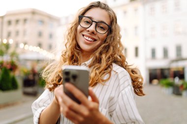 Kıvırcık saçlı ve gözlüklü genç bir kadın gece şehir meydanında akıllı telefonunu incelikle kullanıyor. Arkadaşlarla iletişim. Teknoloji, bağlantı, blog yazma, modern yaşam tarzı kavramı