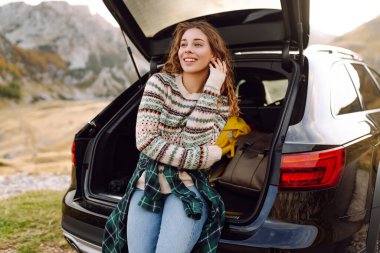 Güneşli bir günde, arabasının açık bagajında güneşli bir arazide oturan neşeli genç bir kadın açık havanın tadını çıkarıyor. Arabayla yolculuk.. 