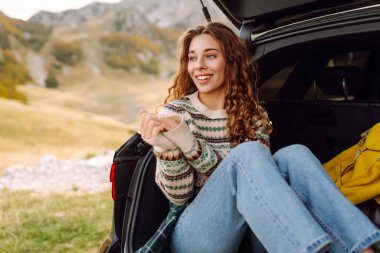 Güneşli bir günde, arabasının açık bagajında güneşli bir arazide oturan neşeli genç bir kadın açık havanın tadını çıkarıyor. Arabayla yolculuk.. 