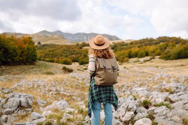 Bir kadın kayalık arazide duruyor, canlı sonbahar ağaçları ve dağlara bakıyor. Çevre turizmi. Etkin yaşam biçimi.