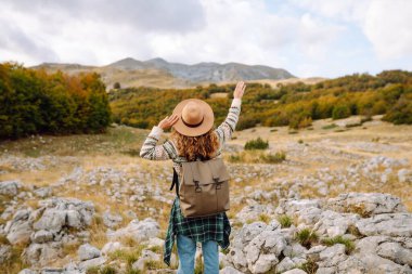 Bir kadın kayalık arazide duruyor, canlı sonbahar ağaçları ve dağlara bakıyor. Çevre turizmi. Etkin yaşam biçimi.