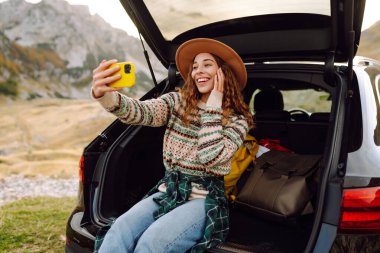 Genç gezgin kadın gün batımında arabasıyla dağlarda selfie çekiyor şık kıyafetlerini ve neşeli ruhunu sergiliyor. Yaşam tarzı, seyahat, turizm, doğa, aktif yaşam.